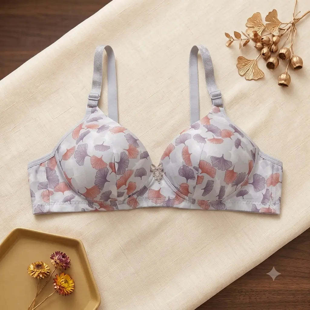 A wireless underwire bra with a light gray and white background and a delicate pink and purple ginkgo leaf pattern, styled on a beige fabric background with decorative gold leaves.
