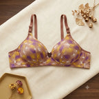A wireless underwire bra with a brown and purple abstract floral pattern, laid flat on a cream-colored fabric background with decorative dried flowers.