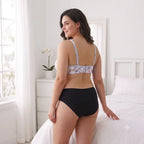 A woman stands in a bright bedroom wearing a two-piece lingerie set. The set includes a black high-waisted brief and a light lavender bralette with a pastel floral pattern, shown from a rear three-quarter angle.