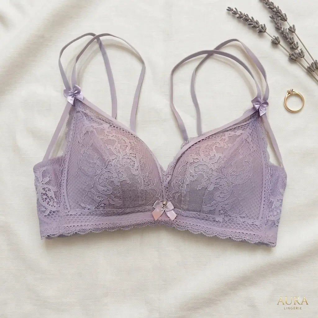 A lavender lace bra with thin adjustable shoulder straps and a small bow at the center, laid flat on a cream fabric background with decorative dried lavender and a ring nearby. The brand name AURA LINGERIE appears in the bottom right corner.
