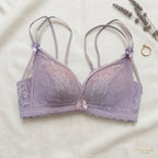 A lavender lace bra with thin adjustable shoulder straps and a small bow at the center, laid flat on a cream fabric background with decorative dried lavender and a ring nearby. The brand name AURA LINGERIE appears in the bottom right corner.