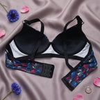 Black underwire bra with a white center panel and blue, pink, and black butterfly-patterned straps, laid flat on a mauve satin background with flower petals and a ring.