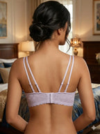 A woman shown from the back wearing a light lavender lace bra with thin adjustable shoulder straps and a lace band across the lower back, photographed in a softly lit bedroom.
