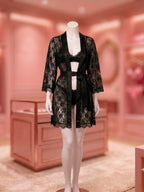A mannequin wearing a black lace robe over a matching black lingerie set, displayed in a softly lit pink boutique interior.
