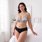 A woman models a two-piece lingerie set in a bright bedroom setting. The set includes a floral-patterned underwire bra with gray straps and black high-waisted briefs.