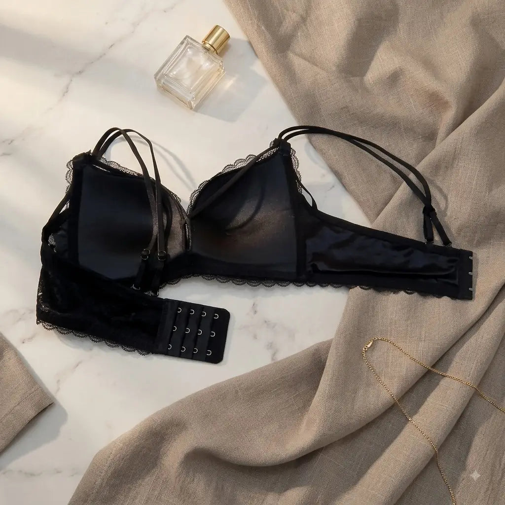 A black lace-trimmed bralette with underwire and adjustable hook-and-eye back, styled on a white marble surface with beige fabric, a perfume bottle, and a gold necklace.