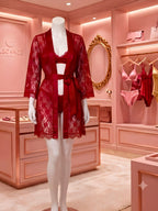 A mannequin in a pink lingerie boutique displays a deep red lace robe over matching red lace underwear. The boutique interior features pink walls, wooden cabinets, a gold mirror, and other pink lingerie items hanging in the background.