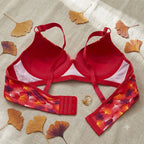 A red bra with floral-patterned underwire cups and a matching red underband, laid flat on a light beige fabric background with scattered dried ginkgo leaves and a small gold ring.