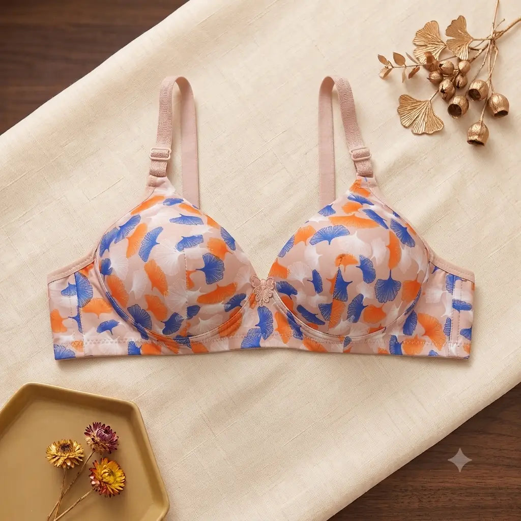 A women's underwire bra with a beige base and an abstract brushstroke pattern in orange and blue, styled flat-lay on a beige fabric background with decorative gold botanical elements.