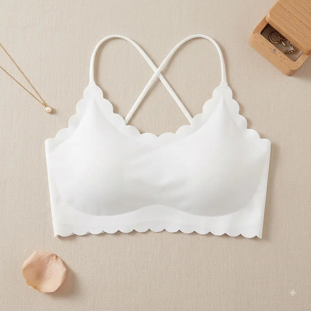 A white wireless bra with scalloped edges and thin crossed back straps, laid flat on a beige textured surface with small decorative props including a necklace, a wooden jewelry box, and a rose petal.