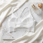 White seamless underwire bra with scalloped edges and crossover straps, styled in a flat lay on a white marble board with draped satin fabric, a gold necklace, a perfume bottle, and a dried rose.