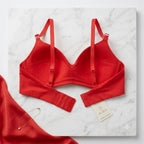 A red wireless bra laid flat on a white marble board, with matching red fabric and a small gold necklace beside it. A paper tag reads AURA.