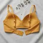 A mustard yellow wireless bra laid flat on a light gray fabric background, with decorative dried leaves and a silver necklace placed around it. The bra has adjustable straps and a triple-hook closure on the underband.