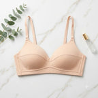 A light peach-beige wireless bra laid flat on a white marble surface, styled with a sprig of eucalyptus leaves on the left and a small clear perfume bottle with a gold cap on the right.