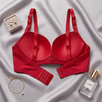 A red seamless underwire bra is laid flat on a draped silver satin fabric, styled with a small jewelry box, a gold bracelet, and a perfume bottle labeled LACE.