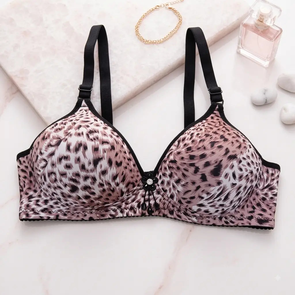 Pink leopard print underwire bra with black trim and adjustable straps, styled on a light marble surface with a gold chain, perfume bottle, and small stones.