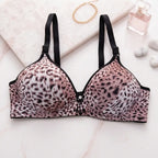 Pink leopard print underwire bra with black trim and adjustable straps, styled on a light marble surface with a gold chain, perfume bottle, and small stones.