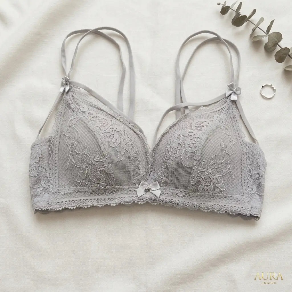A light gray lace bra with thin adjustable shoulder straps and a small bow at the center, laid flat on a white fabric background with decorative eucalyptus leaves and a ring nearby. The brand name AURA LINGERIE appears in the bottom right corner.