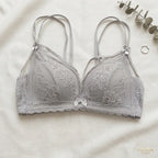 A light gray lace bra with thin adjustable shoulder straps and a small bow at the center, laid flat on a white fabric background with decorative eucalyptus leaves and a ring nearby. The brand name AURA LINGERIE appears in the bottom right corner.