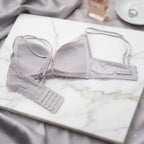 A light gray lace-trimmed underwire bra laid flat on a white marble slab with gray satin fabric and small styling props in the background.