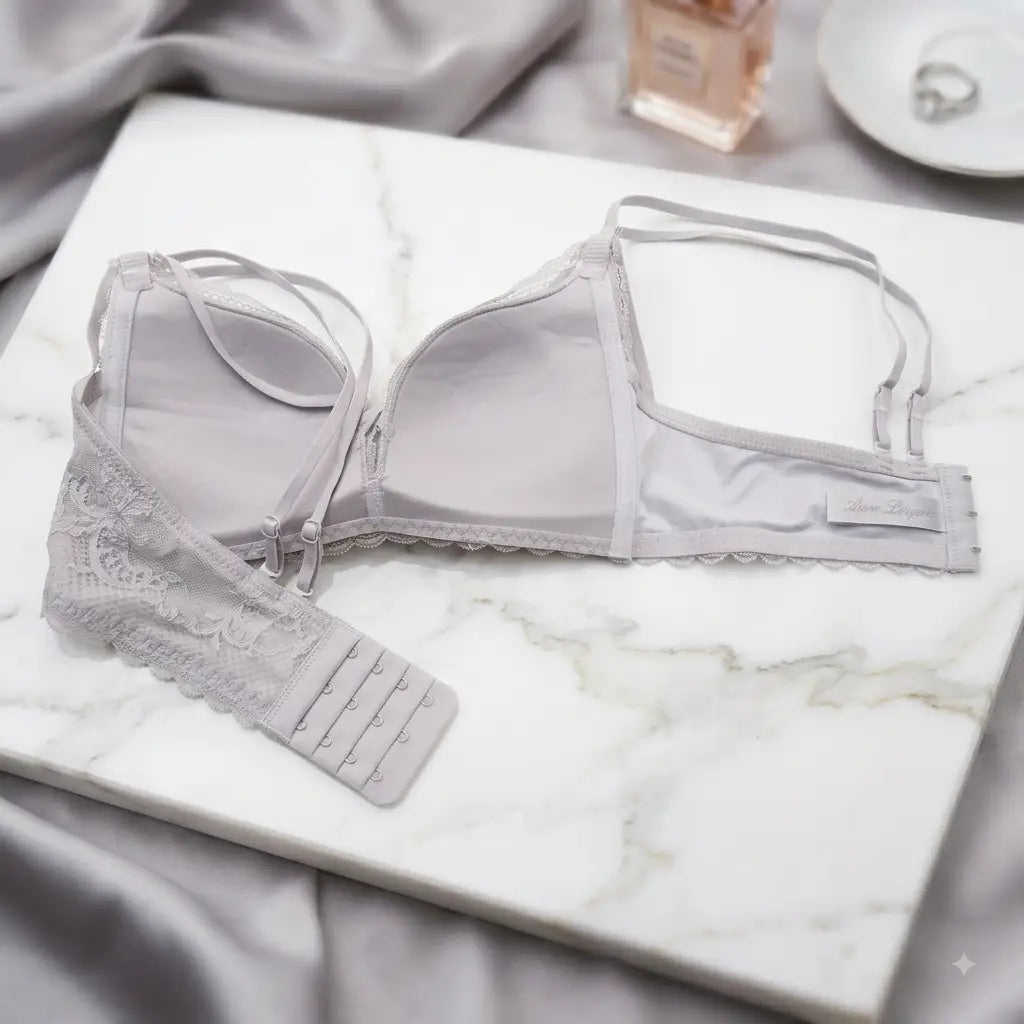 A light gray lace-trimmed underwire bra laid flat on a white marble slab with gray satin fabric and small styling props in the background.