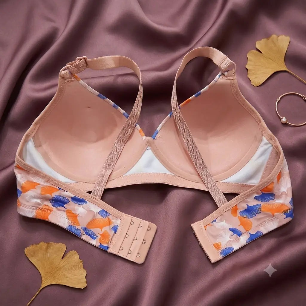 A women's wireless bra laid flat on a mauve satin fabric background, decorated with gold ginkgo leaves and a small gold ring. The bra features a light peach-beige color with a blue and orange floral pattern on the underwired cups and straps.