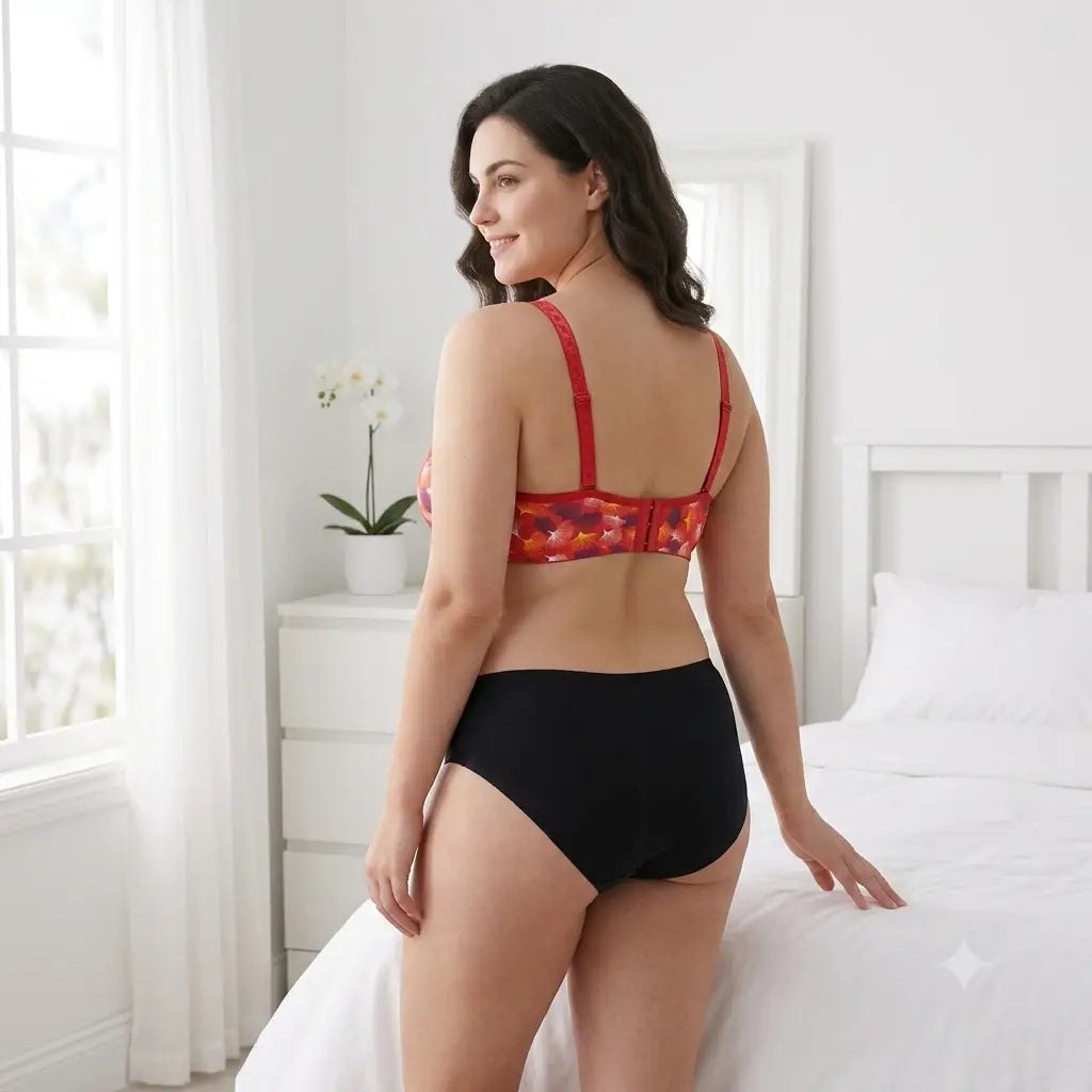 A woman stands on a white bed in a bright bedroom, wearing a two-piece lingerie set. The set features a red underwire bra with a red, orange, and yellow floral pattern, paired with high-waisted black briefs.