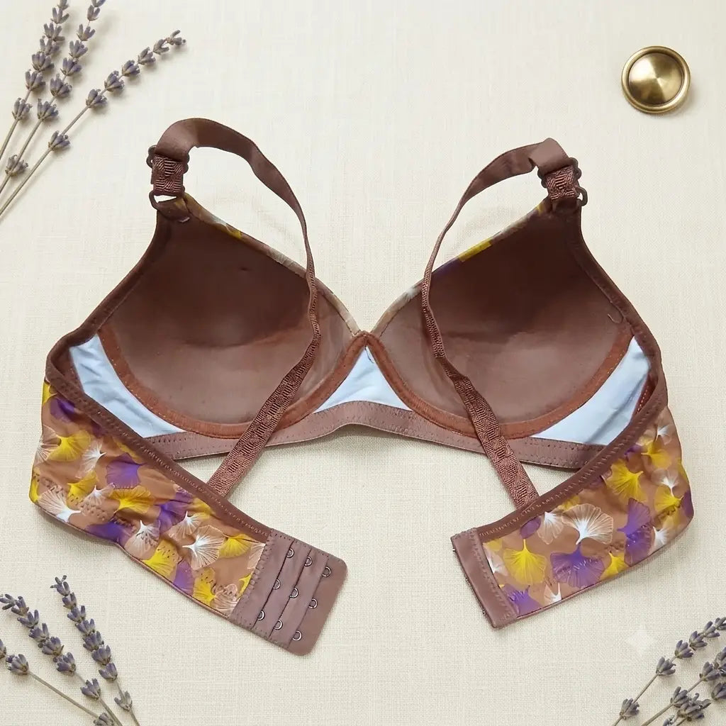 A brown lace and mesh bra with a floral patterned underwire band, laid flat on a textured cream fabric background with decorative lavender sprigs and a gold coin.