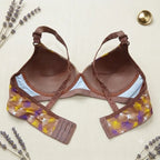 A brown lace and mesh bra with a floral patterned underwire band, laid flat on a textured cream fabric background with decorative lavender sprigs and a gold coin.