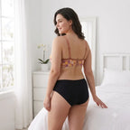 A woman stands on a white bed in a bright bedroom, wearing a floral bralette with matching bra straps and black high-waisted briefs, shown from the back.