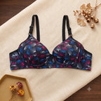 A wireless underwire bra with a dark navy and black base, decorated with a colorful abstract leaf pattern in blue, pink, and purple. The bra is laid flat on a beige fabric background with decorative dried botanical elements.