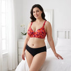 A woman modeling a two-piece lingerie set in a bright bedroom. The set includes a red and orange abstract patterned bra and solid black high-waisted briefs.