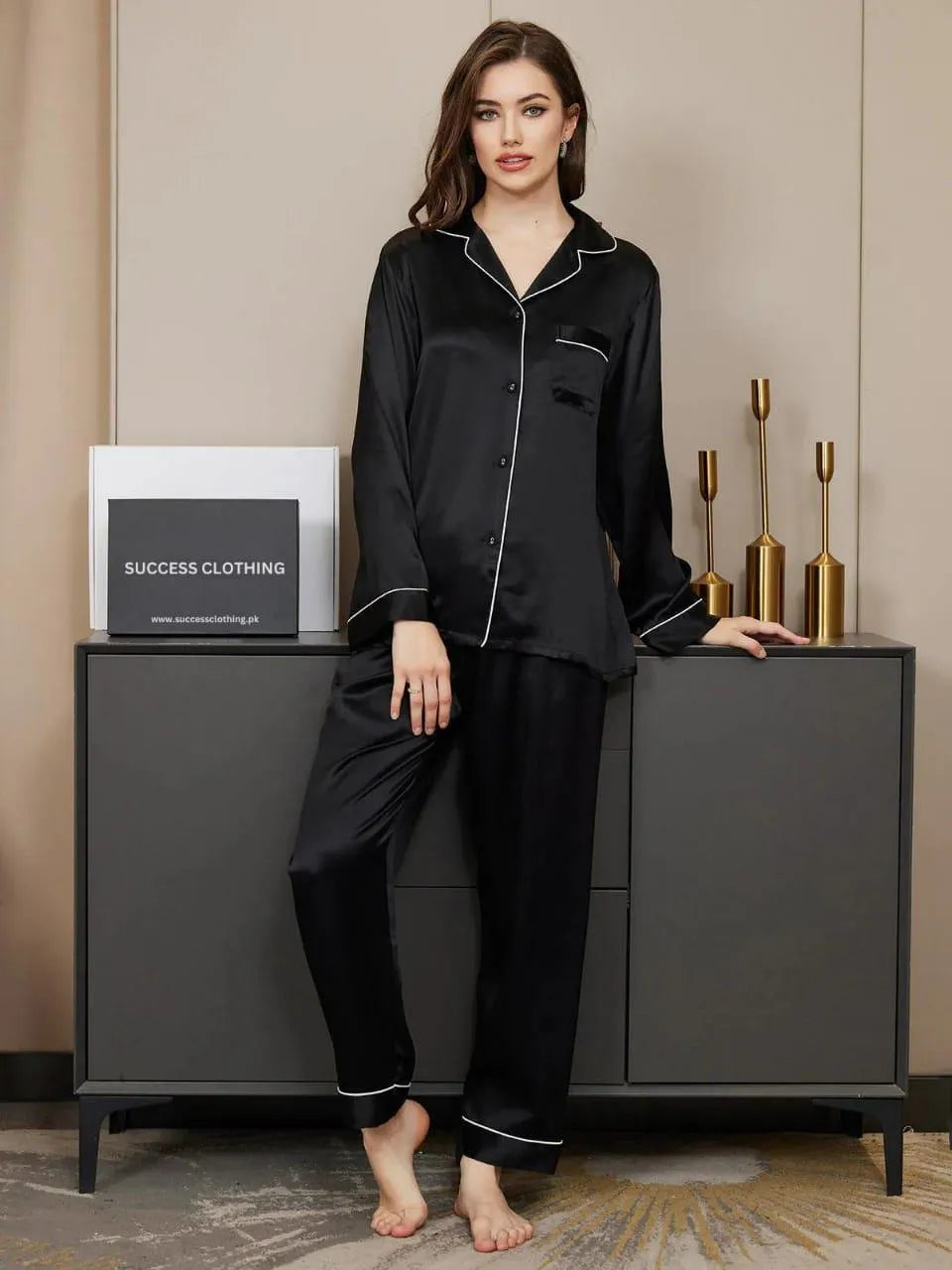 Woman wearing black satin pajamas standing next to a cabinet with decorative items.