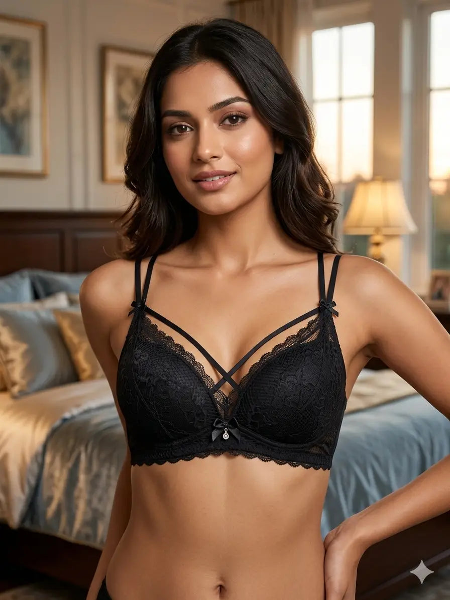A woman modeling a black lace bra in a softly lit bedroom setting. The bra features thin adjustable straps, delicate lace cups, and a small bow detail at the center front.