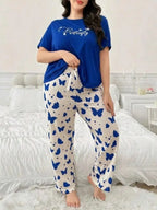 A woman modeling a two-piece pajama set in a bedroom setting. The set includes a royal blue short-sleeve t-shirt with 'Butterfly' printed in white script on the chest, and matching cream-colored pants covered in a blue butterfly pattern with a white drawstring waist.