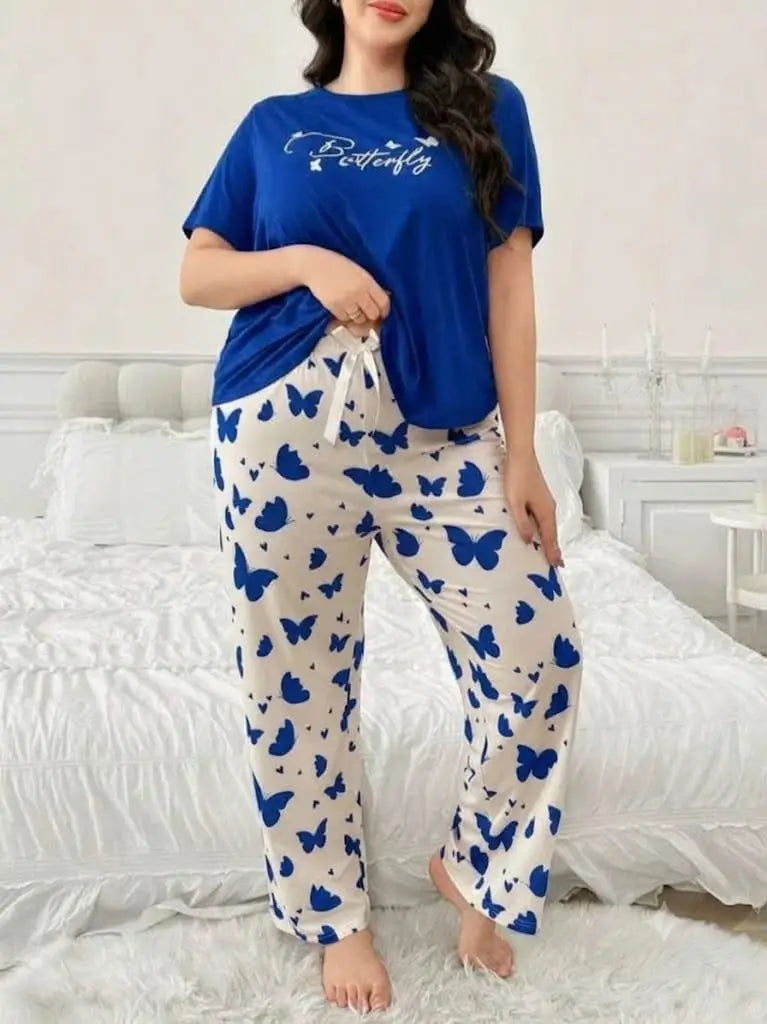 A woman modeling a two-piece pajama set in a bedroom setting. The set includes a royal blue short-sleeve t-shirt with 'Butterfly' printed in white script on the chest, and matching cream-colored pants covered in a blue butterfly pattern with a white drawstring waist.