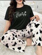 A woman wearing a matching pajama set consisting of a black short-sleeve t-shirt with 'Butterfly' written in white script and small white butterflies, paired with white pants featuring a black butterfly and heart pattern. She is sitting on a white faux fur rug in a bedroom setting.