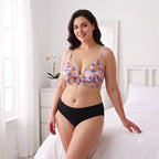 A woman models a two-piece lingerie set in a bright bedroom. The set features a multi-colored floral pattern bra in orange, purple, and white, paired with solid black high-waisted briefs.