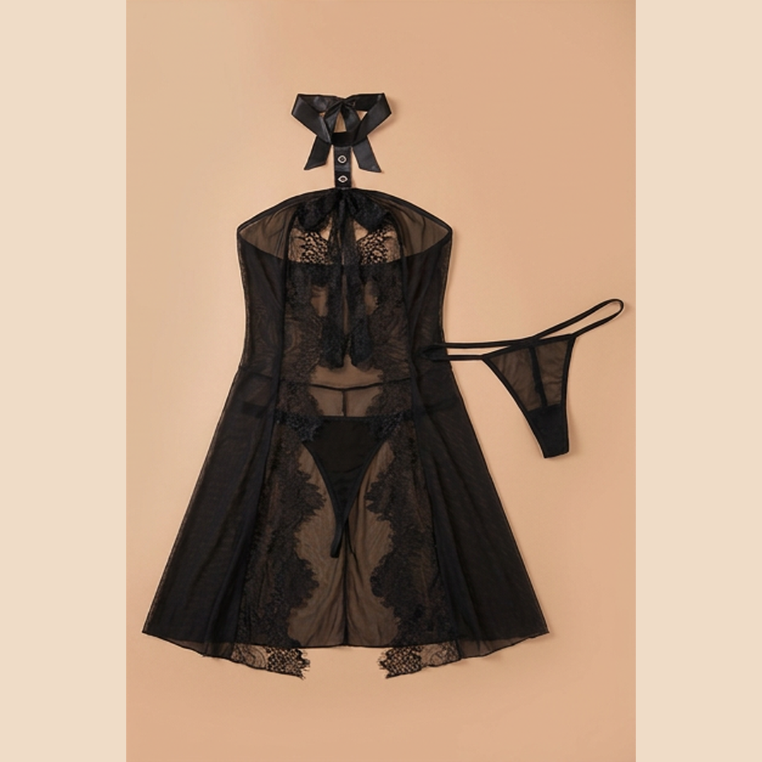 Black sheer lace lingerie set featuring a halter-neck chemise with a tie at the neck, a matching thong, and a sheer overlay skirt with floral lace trim, displayed on a warm beige background.
